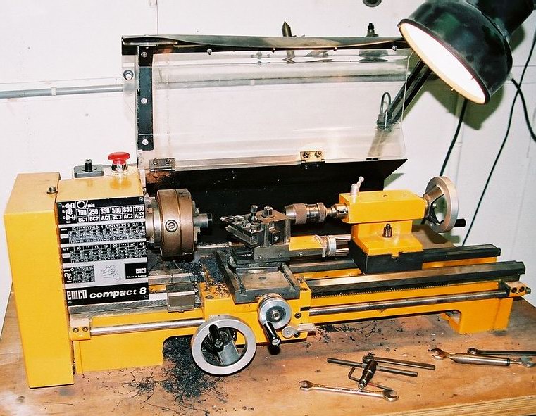 Yellow conventional metal lathe with a chuck, toolpost, tailstock, transparent guard and task lamp.