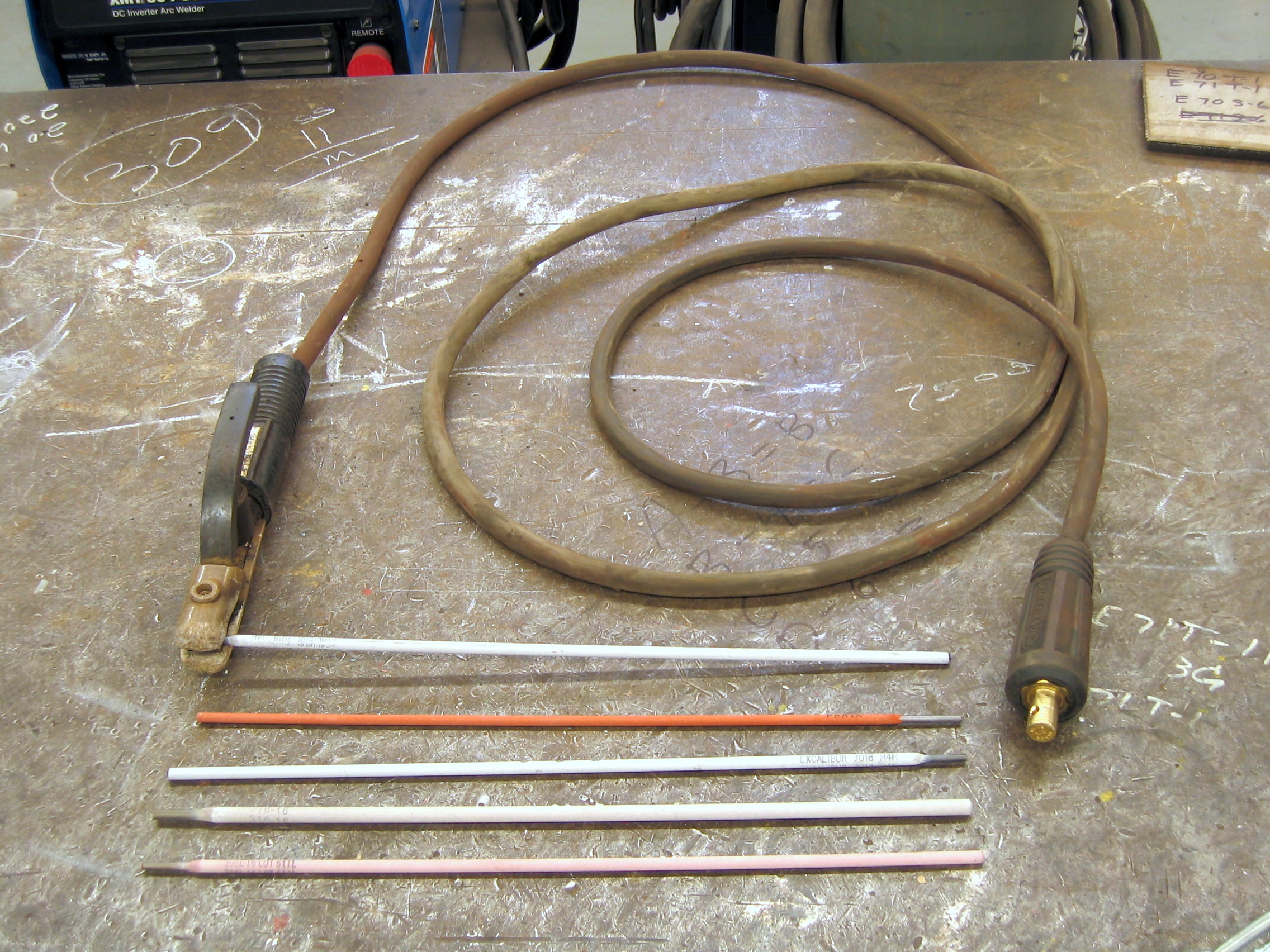 Electrode holder and several coated stick welding electrodes on a steel shop bench; contextual photograph, not identified E7016.